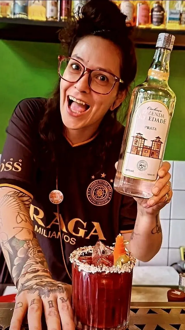 Prix atrás do balcão, rindo, com uma garrafa de cachaça e um coquetel